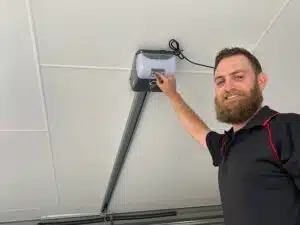 A garage door technician is smiling at the camera, with his hand on a garage door opener